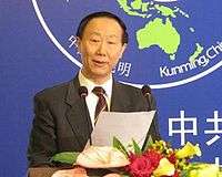 a serious looking nearly bald man, wearing a suit and a dark coloured tie while holding a piece of paper