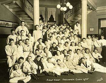 About 50 members of the 1935 freshman class gather on the first floor of their dormitory, which has columns, a chandelier with spherical lights, and multiple staircases.