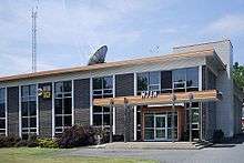 A two-story, dark- and white-brick building with tall, dark windows. On its flat roof is a white satellite dish and in the background is seen a tall radio tower. Over the entrance are the letters "WTEN".