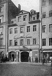 A building with four main storeys with an open shop to one side of an arched entrance and garret windows in the roof. A sculpted figure of an animal is above the arch.