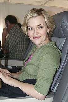 A woman with short hair in an airplane seat wearing a green sweatshirt and smiling