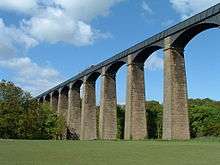 Looking up toward a metal aqueduct, supported by multiple tall, stone pillars and arches.