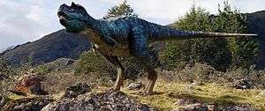 The Gorgosaurus, a bipedal dinosaur depicted with iridescent scales, stands on mostly rocky terrain with deciduous trees behind it.