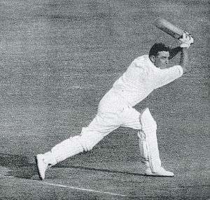 A dark haired man wearing a white shirt, white trousers and cricket pads, holds a cricket bat in the air having just played a shot.