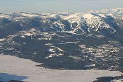 Aerial view of a forested mountainous area, with ski trails.  In the foreground is a partially frozen body of water.