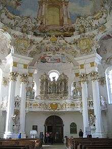 Ornate church interior, looking toward the entrance. The interior is white, the doors flanked by two pairs of columns which stretch to the richly painted ceiling. Above the entrance is the church's pipe organ.