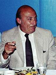 A photograph of an elderly man, seated and wearing a light-gray suit and tie.