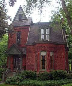 A brick house with a high pointed roof on its left and a lower, trapezoid-shaped one on its right. The first story has a pointed-roof porch on the left and a section sticking out from the brick on its right.