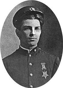 Head and shoulders of a young white man wearing a cap pushed high up on his forehead and a plain military jacket with a star-shaped medal hanging from a ribbon pinned to his left breast.