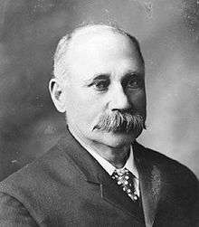 A white man with receding light hair and a thick grey moustache, wearing a suit