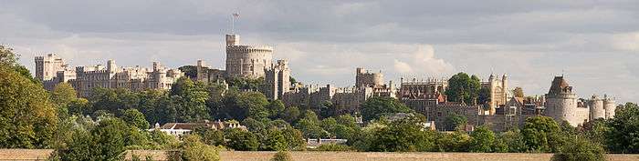 A photograph of a white-grey stone castle, running left to right; trees are in the foreground, with a large white tower the most prominent part of the castle in the middle of the shot.