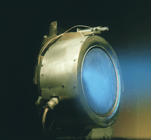 A metal cylinder with electrodes attached to its side. Blue diffuse light is coming out of the tube.