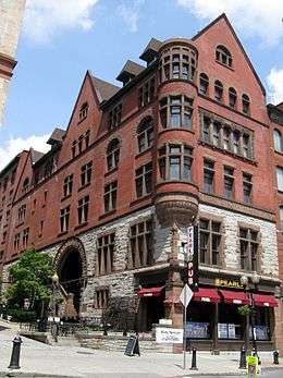 A five-story building with a lower section in stone and the upper section in brick. It has decorated windows, a tower on the corner, and a pointed roof. At the street below it is a glass-faced section with signs identifying it as the Pearl Street Pub.