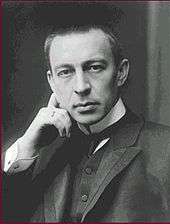 A clean-shaven young man with close-cropped hair, wearing a dark jacket, dress shirt with a high stiff collar and tie, looking toward the viewer with one hand on the side of his head